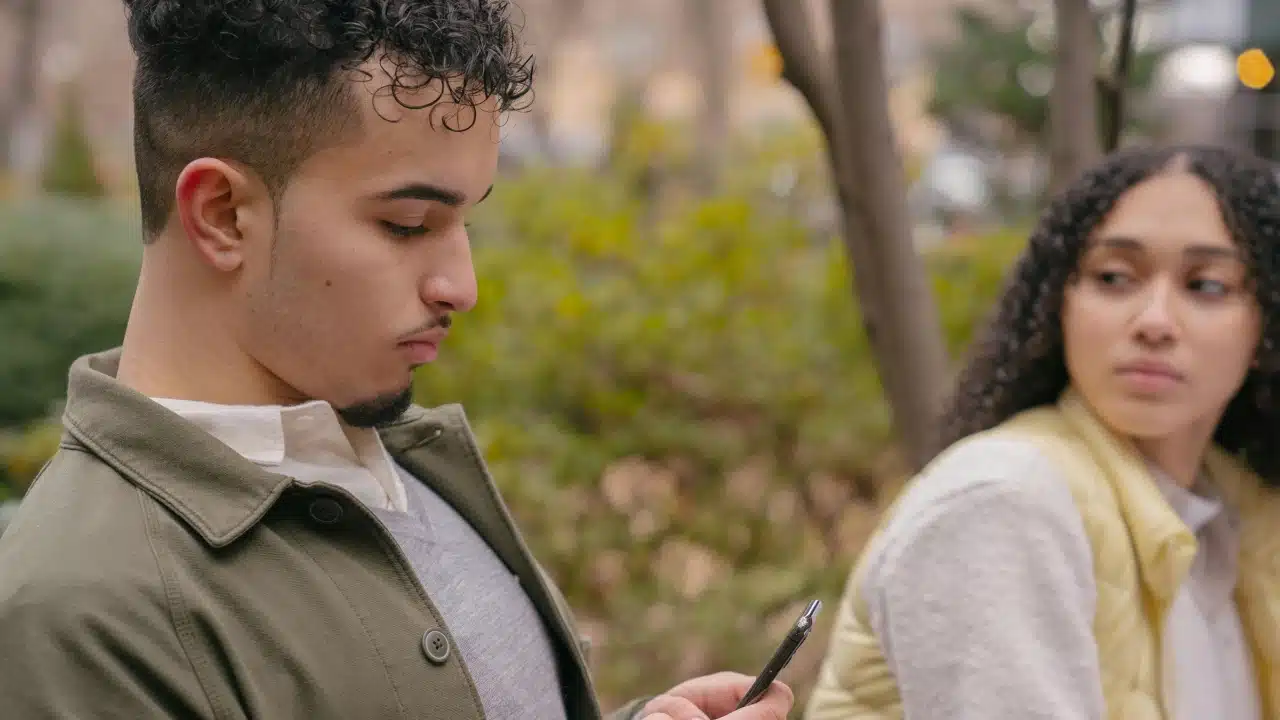 A man looks at a smartphone while a woman behind him watches with a concerned expression.
