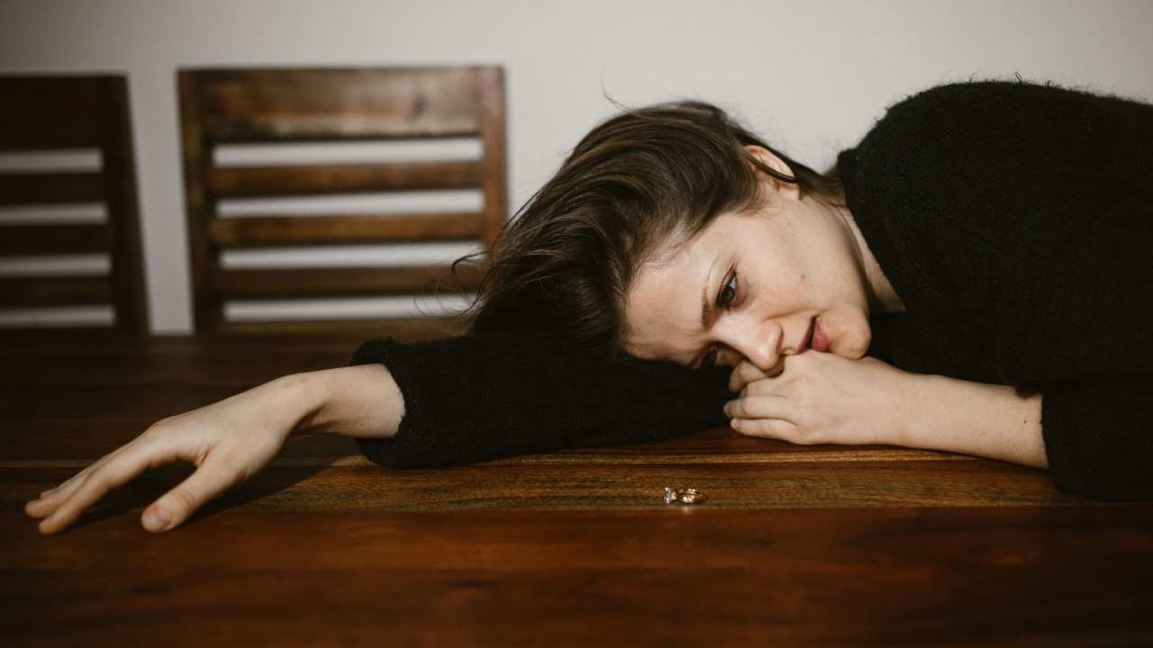 A woman rests her head on a wooden table next to a small gold ring.