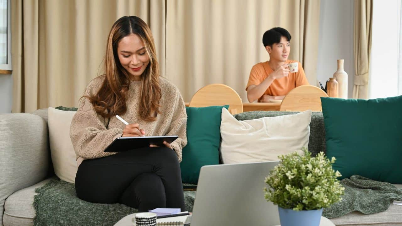 A woman writes on a tablet while a man sits in the background drinking coffee.