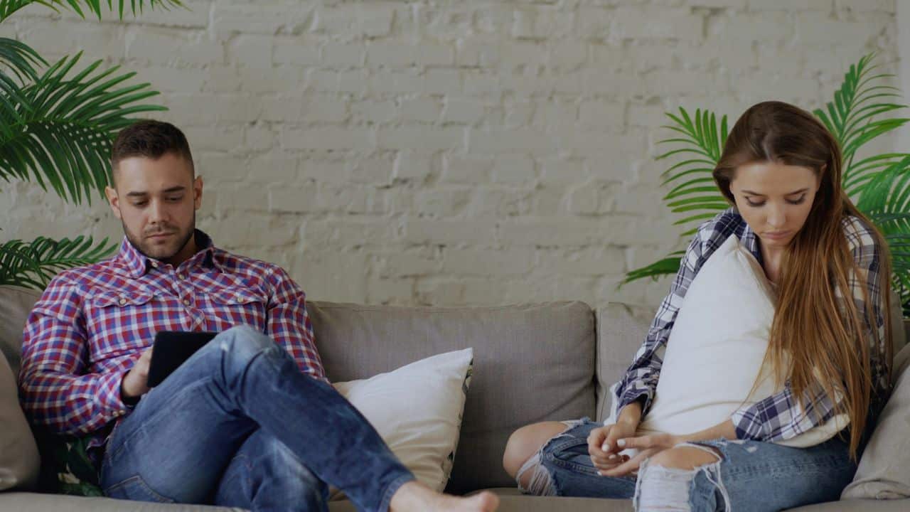 A man and woman sitting apart on a sofa, both looking down at their laps.