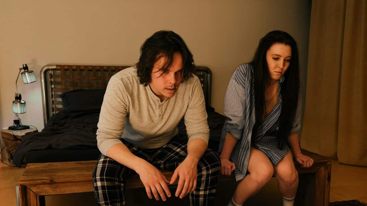 A man and a woman sit on the edge of a wooden bed frame looking down.