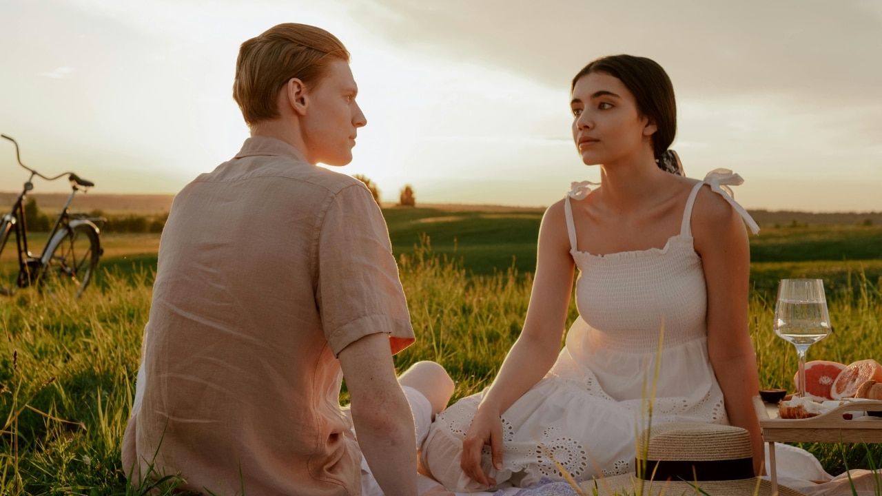 A man and woman sit on the grass in a field during a sunset facing each other.