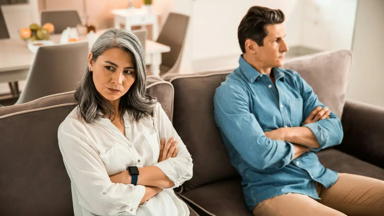 A man and woman sit on a sofa with arms crossed, looking in opposite directions.