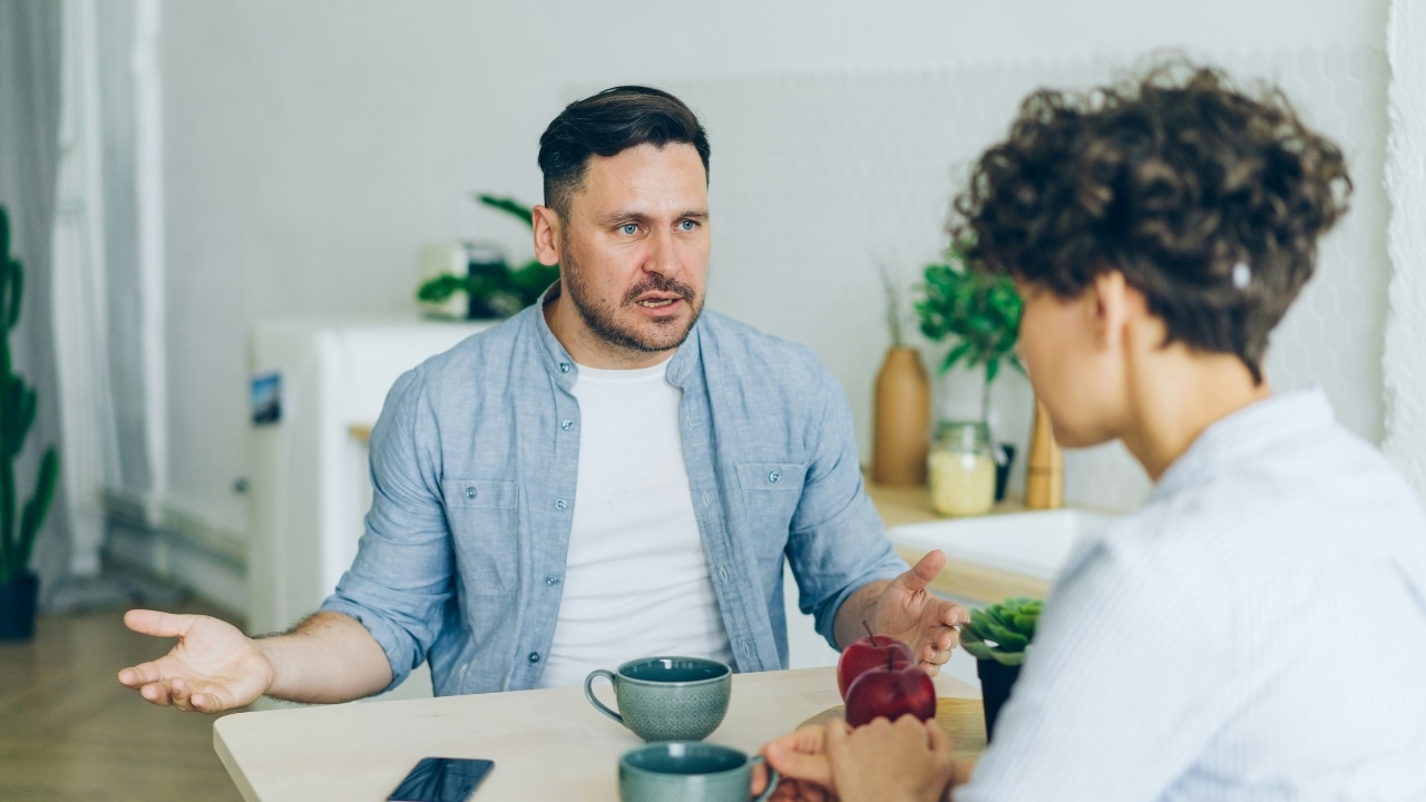 A man gestures with open palms while speaking to a person across a wooden table.