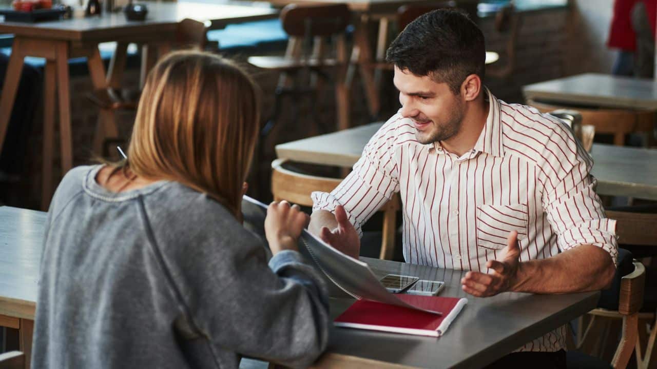 A man in a striped shirt gestures with both hands toward a woman seated across from him at a table.