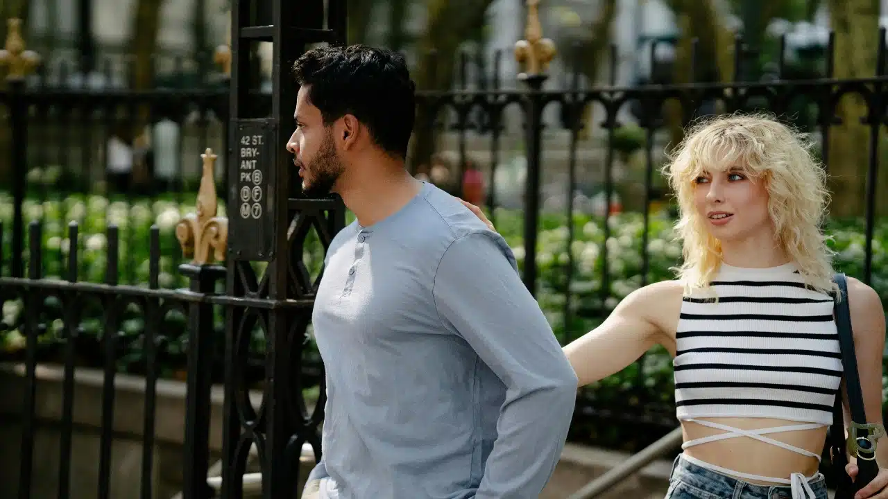 A woman in a striped top reaches out to touch a man’s shoulder near a fence.