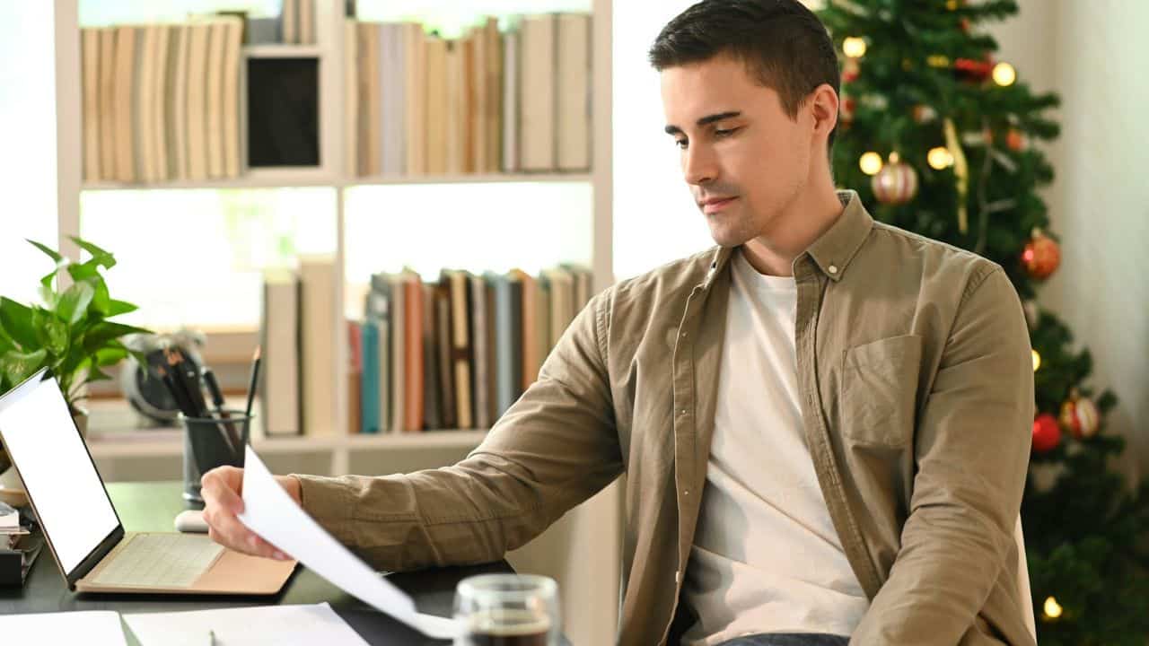 A man sits at a desk holding a piece of paper near a laptop computer.