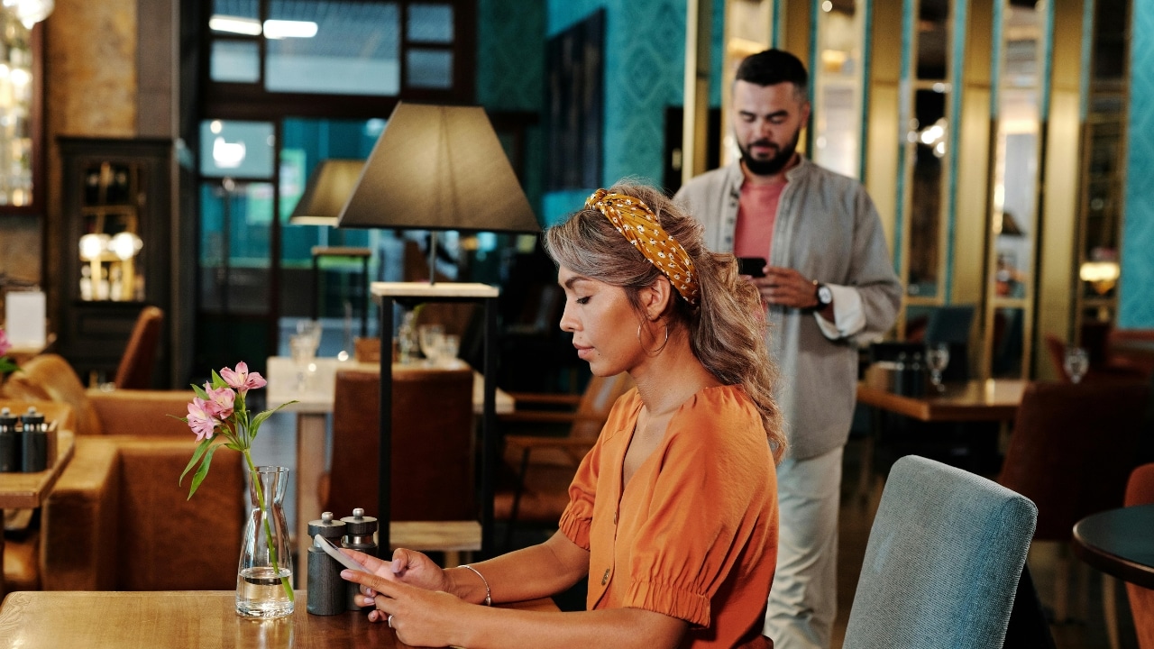 A woman in an orange dress sits at a table while a man stands behind.