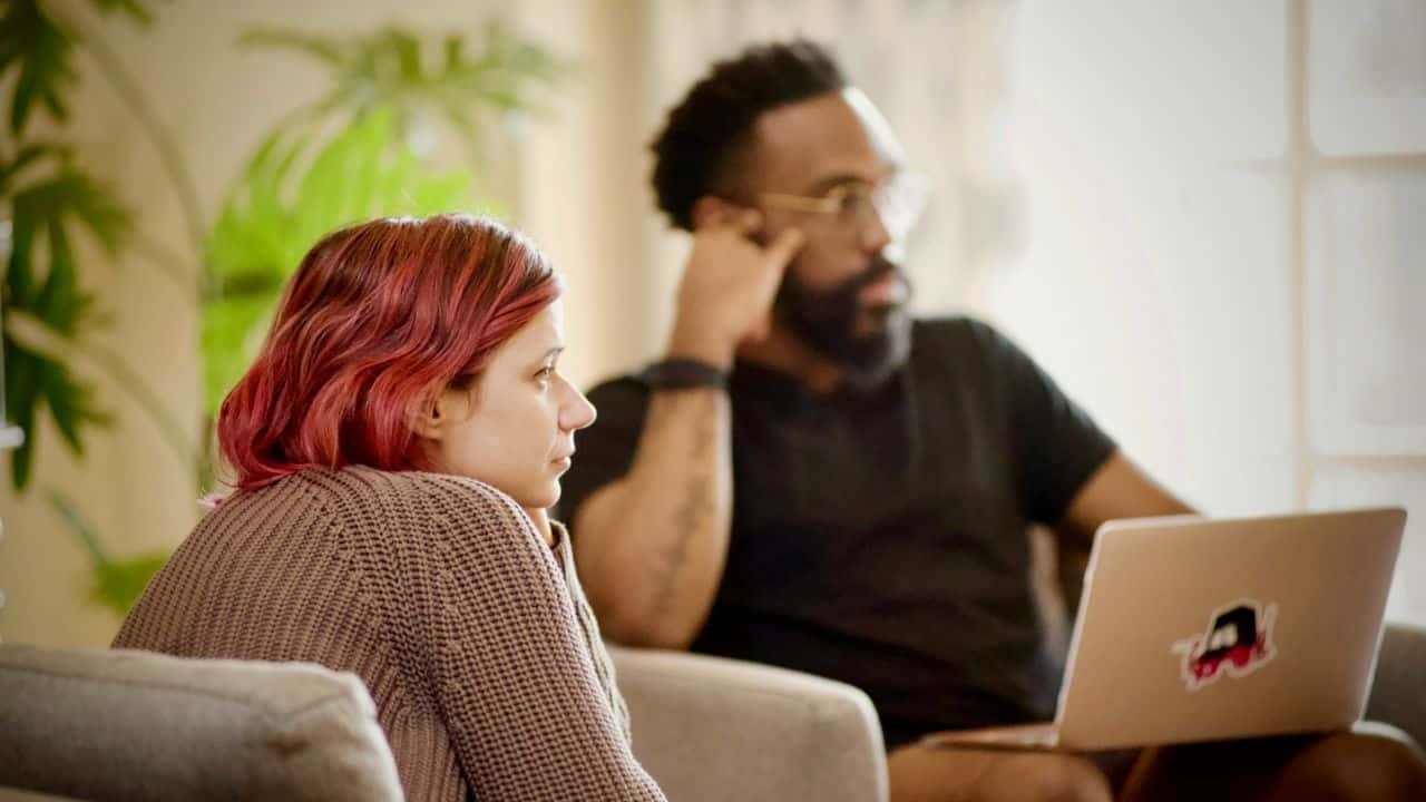 A woman with red hair sits on a couch near a man with a beard and a laptop.