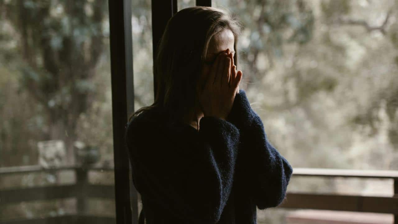 A woman covering her face with her hands while standing by a window.