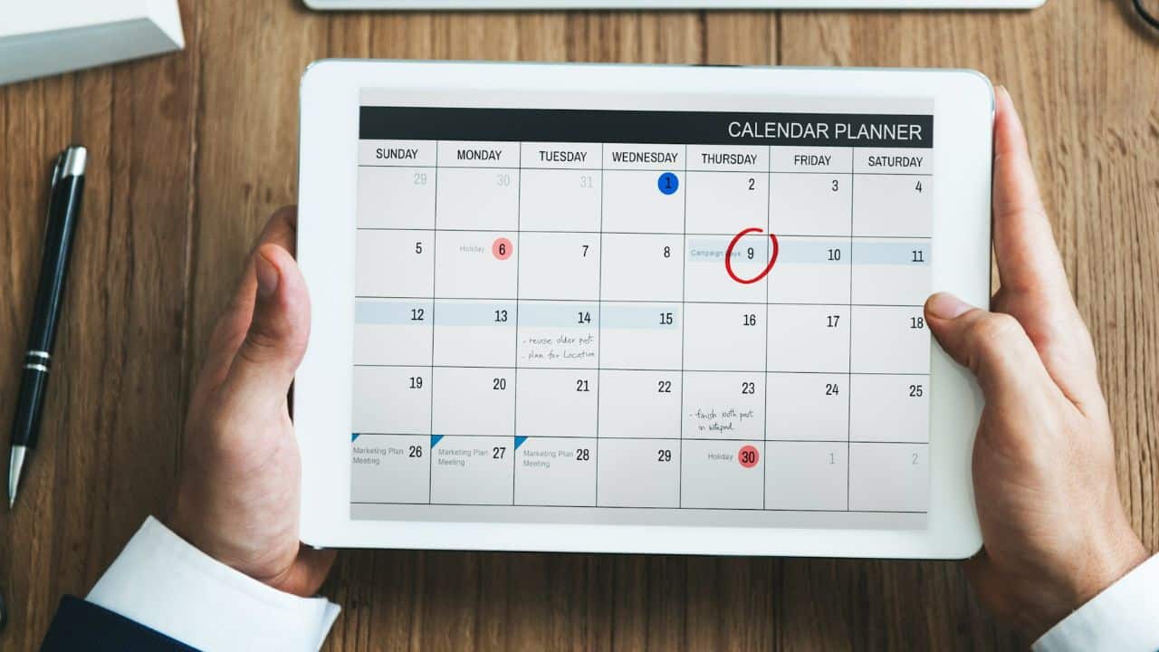Two hands hold a white tablet displaying a digital calendar planner with various dates circled.