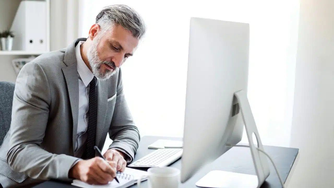 A grey-haired man in a suit writes in a notebook at a bright office desk.