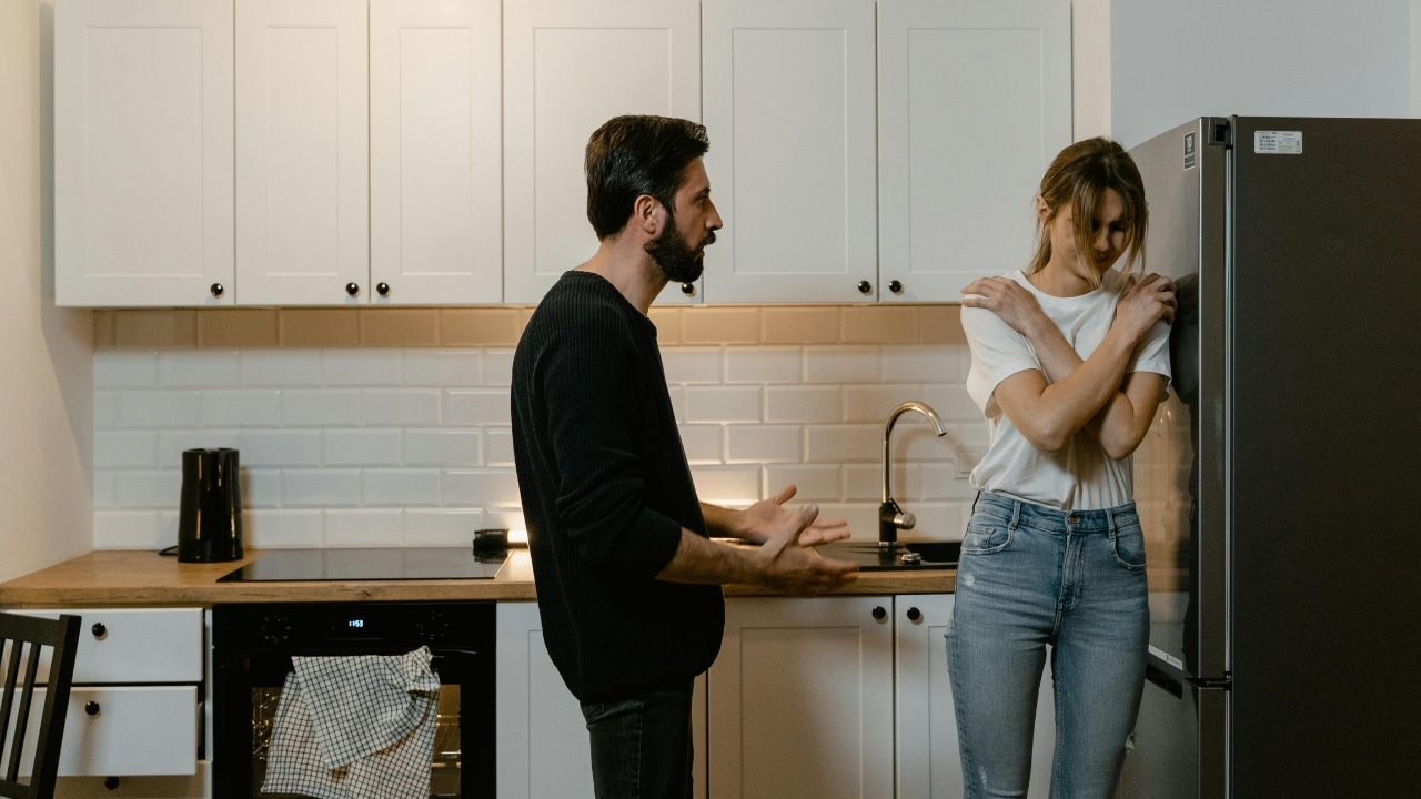 In a kitchen, a man gestures with open hands toward a woman leaning against a refrigerator.
