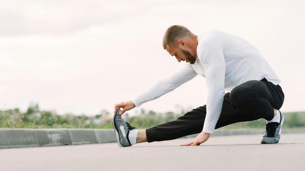 A man in athletic wear perform a leg stretch on a paved path outdoors.