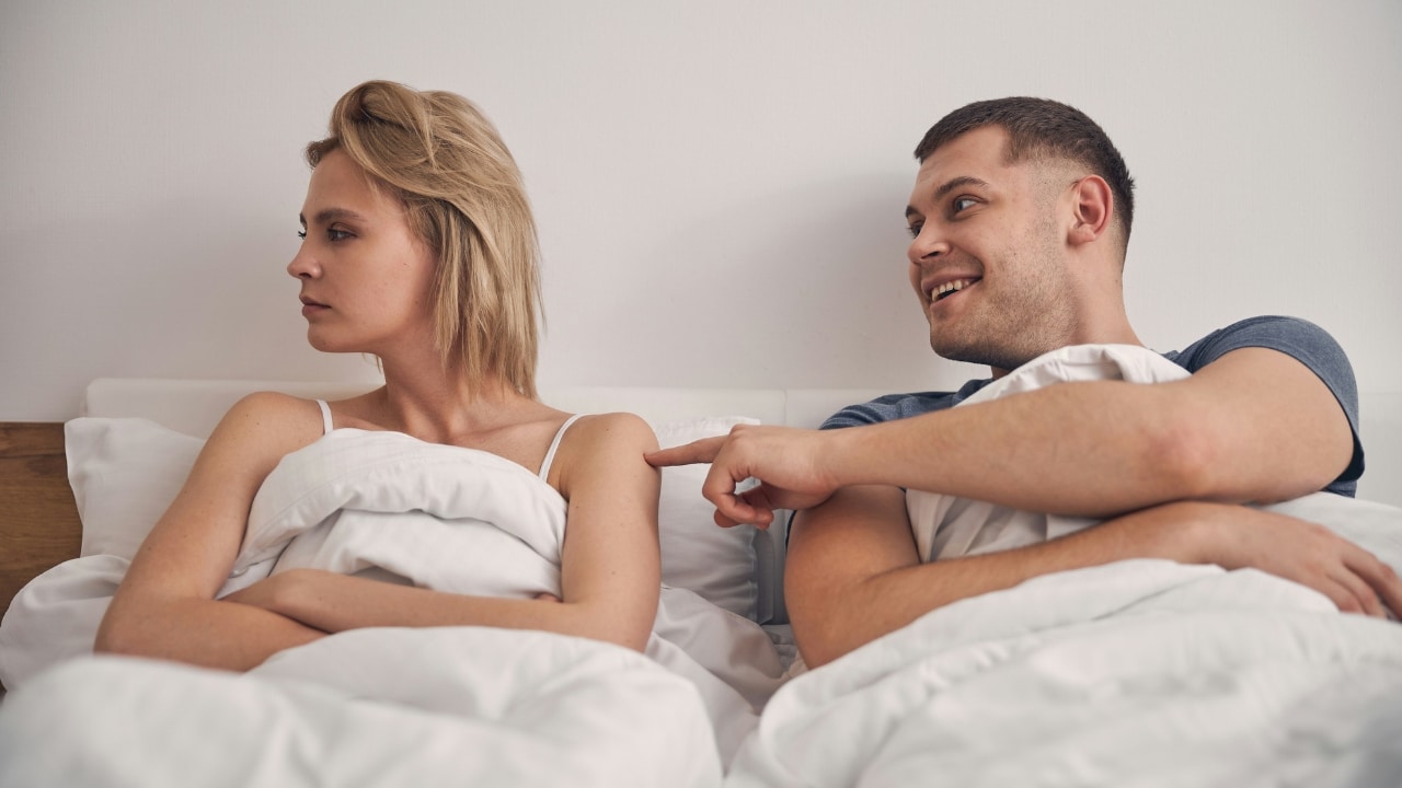A woman sits with crossed arms in bed while a man smiles and points.