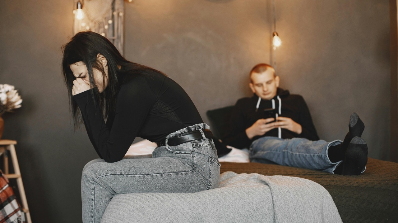 A woman sits on a bed covering her face while a man uses a phone.