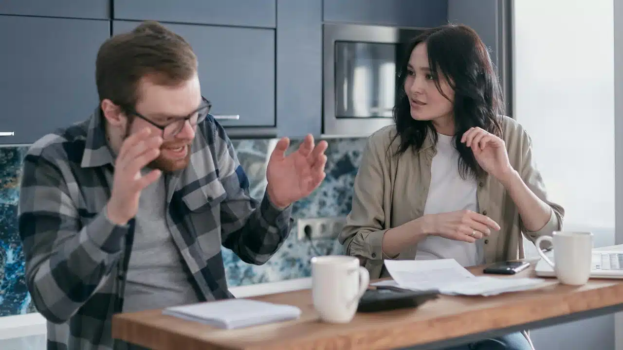 A man gestures with raised hands over papers while a woman watches him from across.