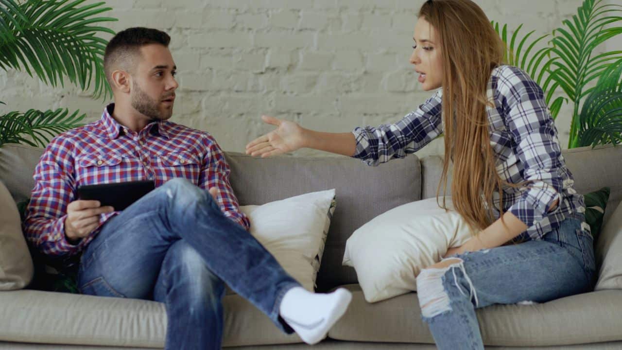 A man holding a tablet sits on a couch while a woman gestures toward him.