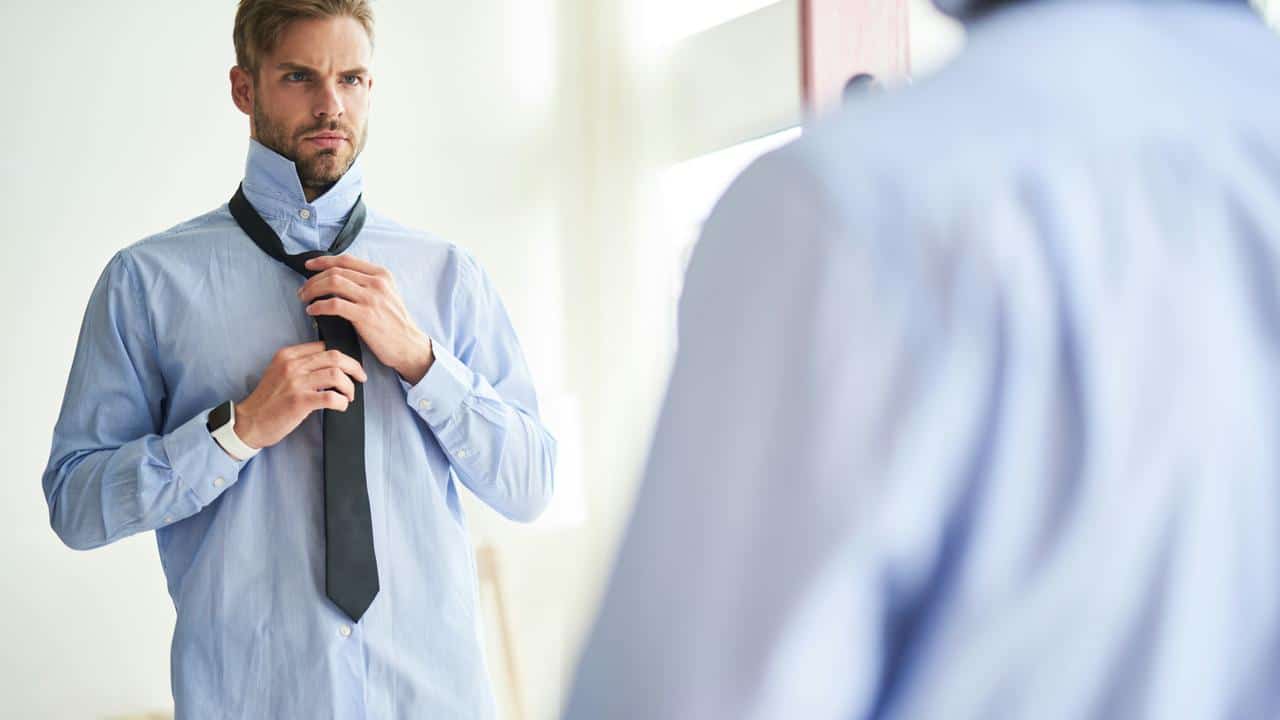 A man in a blue shirt tying a black tie in front of a mirror.