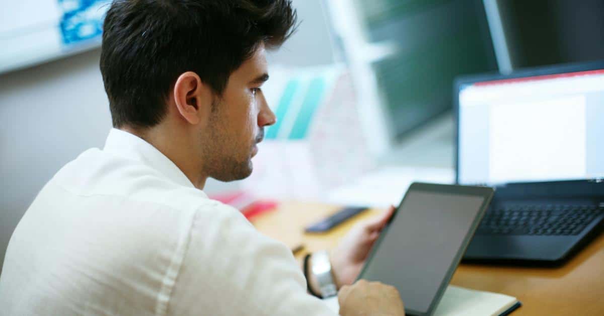 A man focused on a tablet while working at a desk with a laptop.
