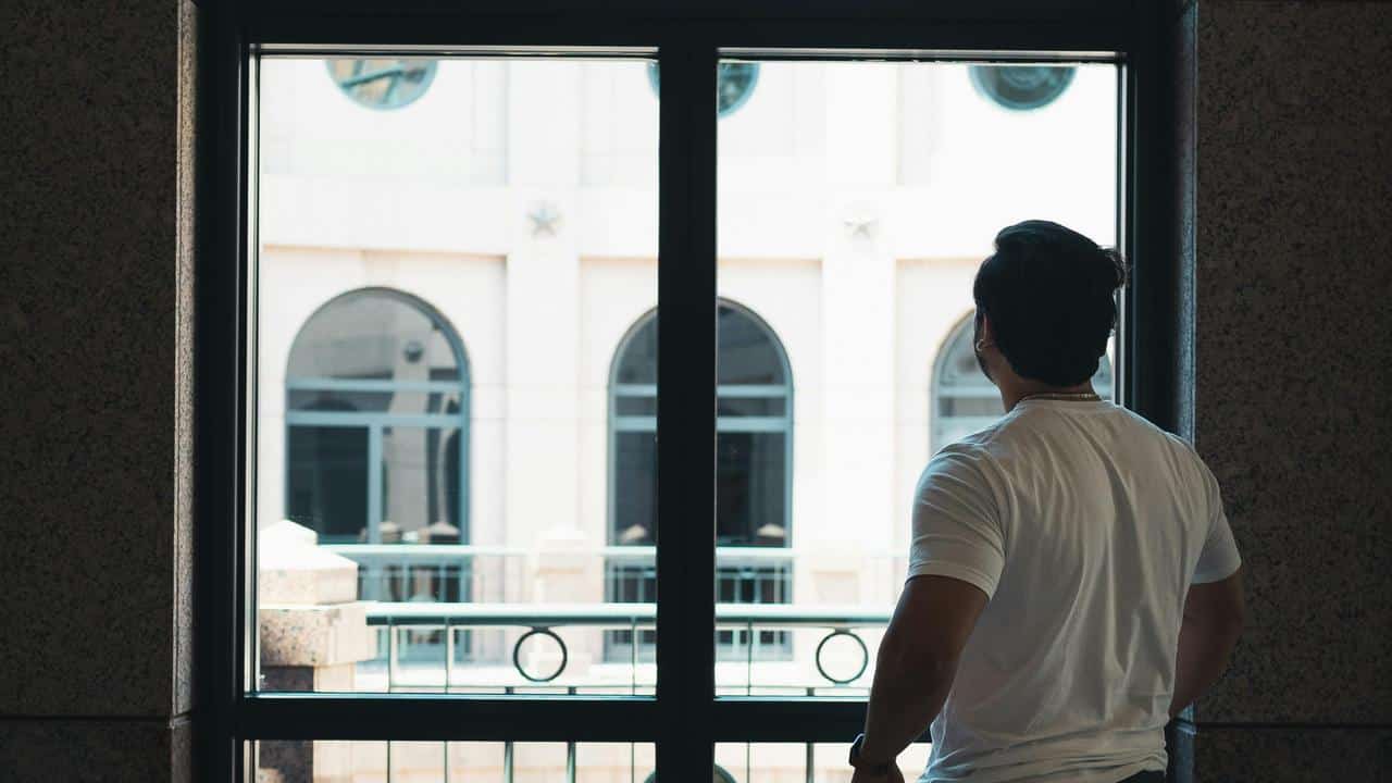A man standing indoors looking out through a large window.