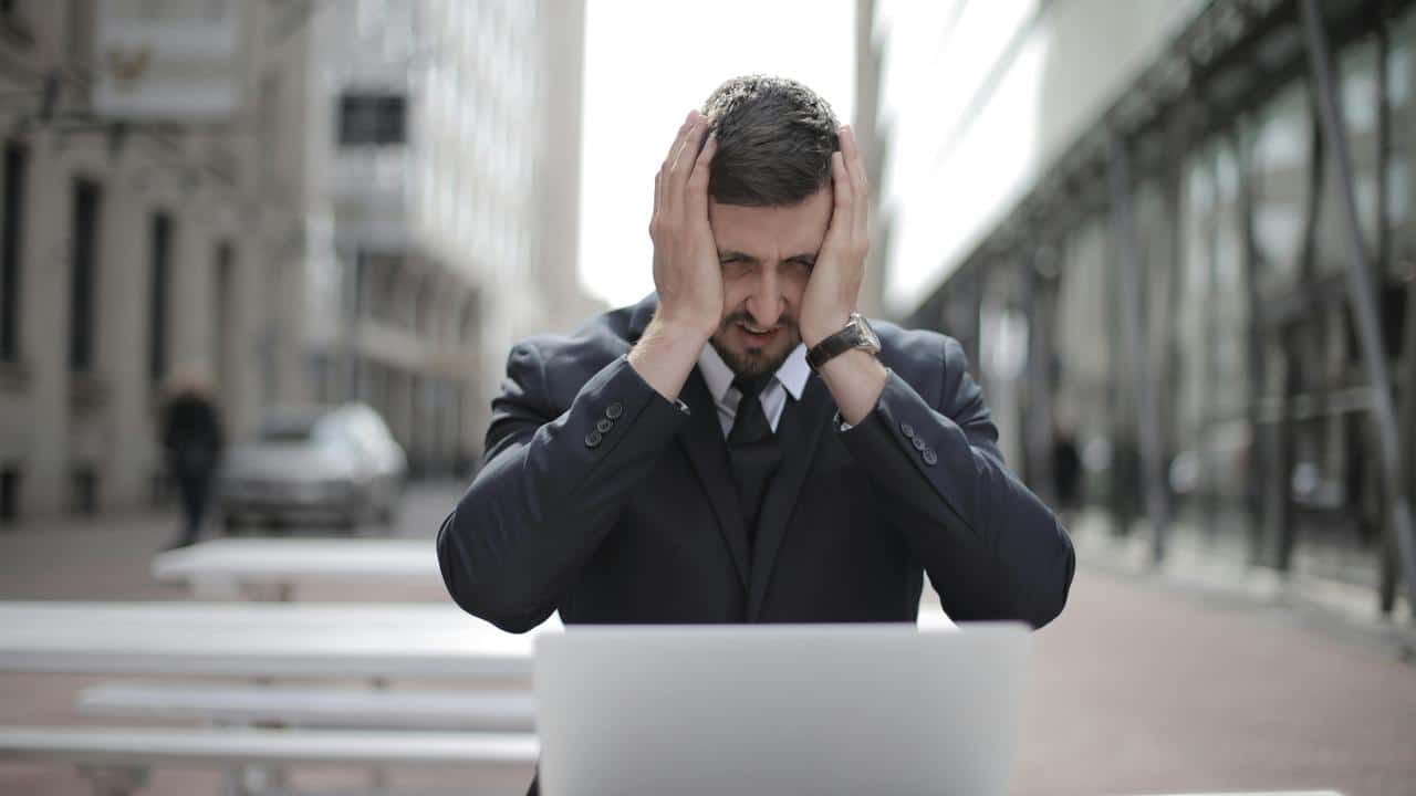 A stressed businessman holding his head while sitting in front of a laptop outdoors.