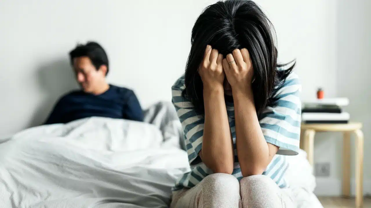 A woman sits with her head in her hands while a man sits behind her in bed.