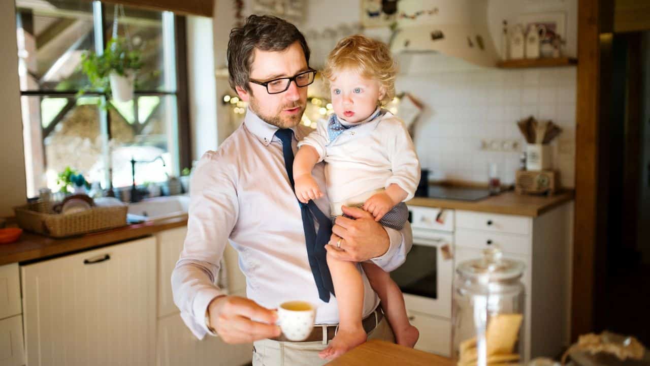 A man in a dress shirt and tie holds a baby and a cup of coffee in the kitchen.