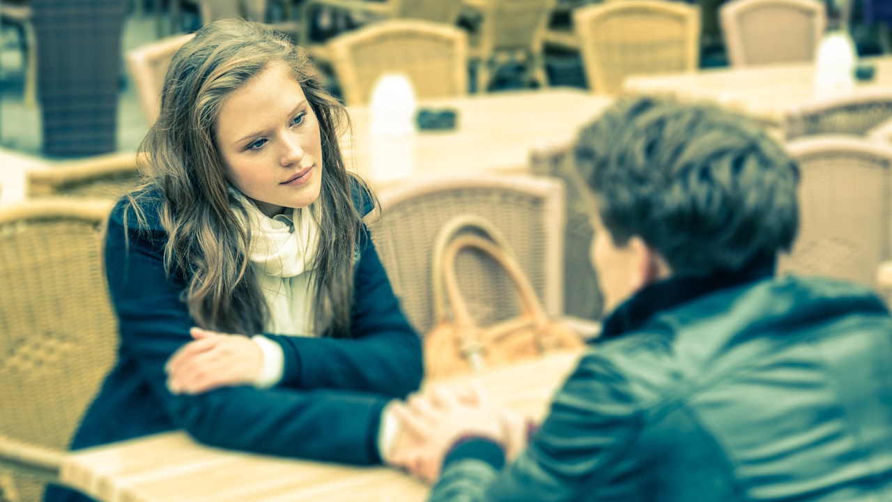 A woman intently listening to a blurred person across a cafe table.