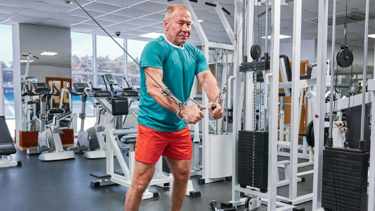 Older man in a teal shirt and red shorts using a cable machine for a chest workout in a gym.