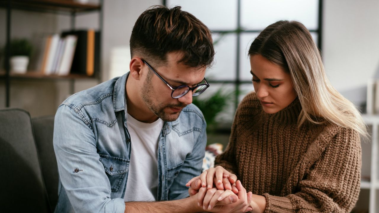 A serious-looking couple holding hands while sitting on a couch in a living room.
