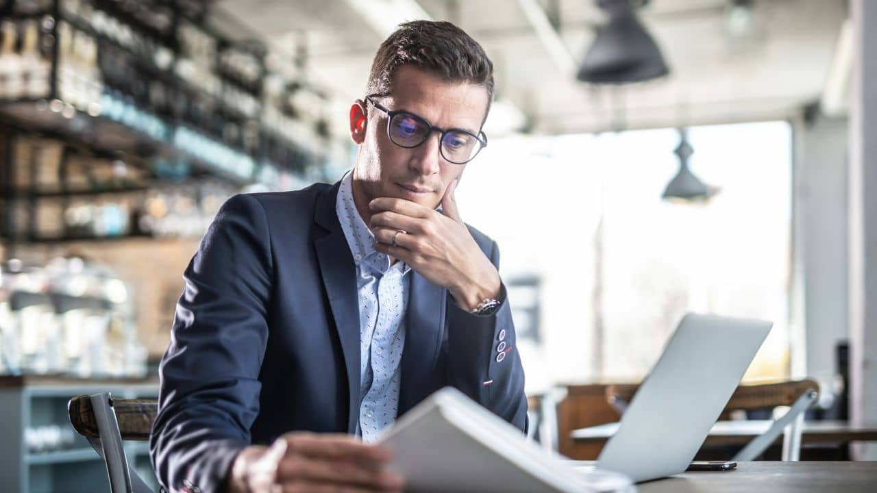 A serious man in a suit and glasses reading a document near a laptop in a cafe.
