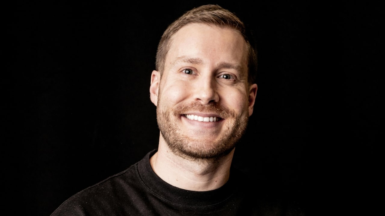 A man with a short beard and brown hair smiles broadly against a black background.