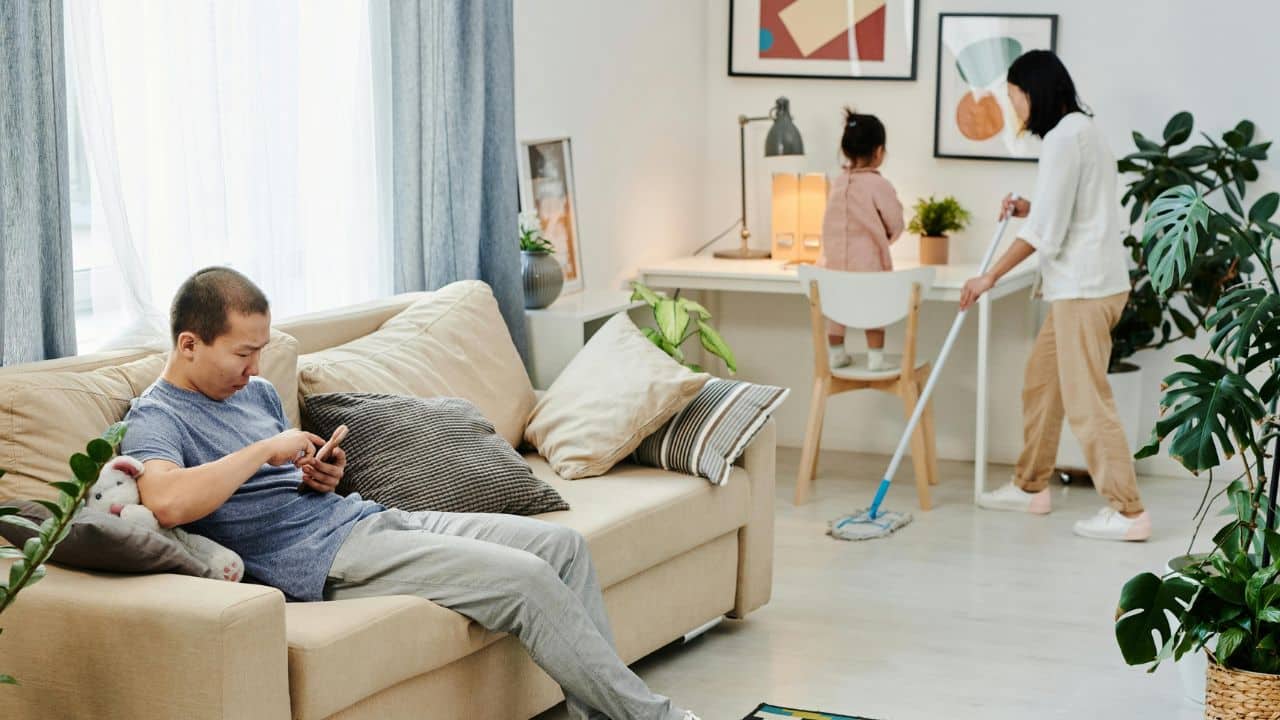 A man uses a phone on a sofa while a woman mops the floor nearby.