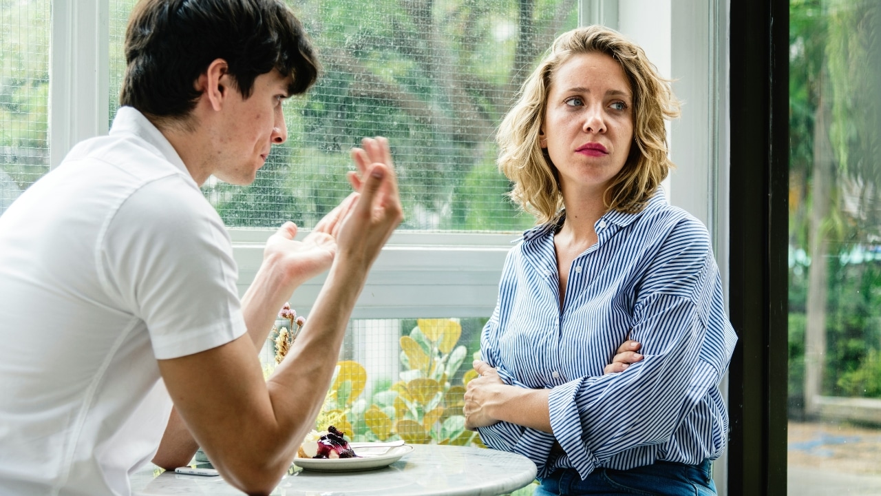 A man gestures with his hands while speaking to a woman looking away with crossed arms.
