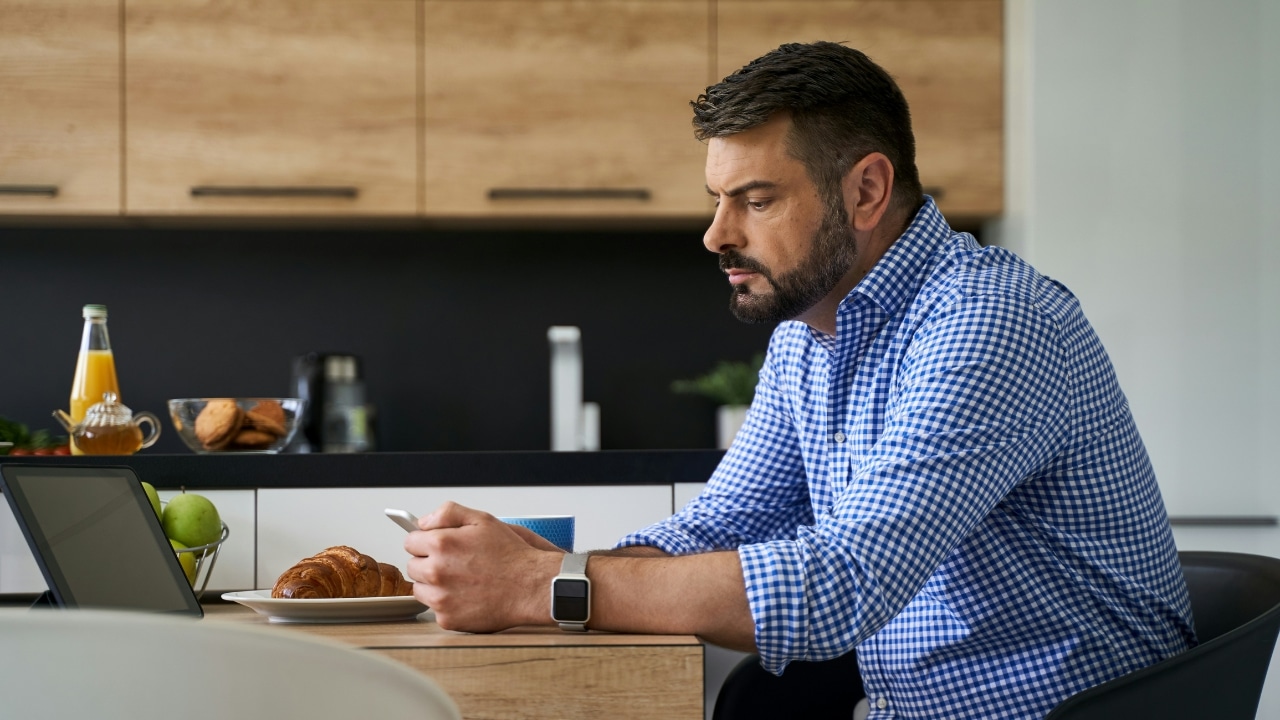 A bearded man in a checkered shirt sits at a kitchen counter looking at a smartphone.