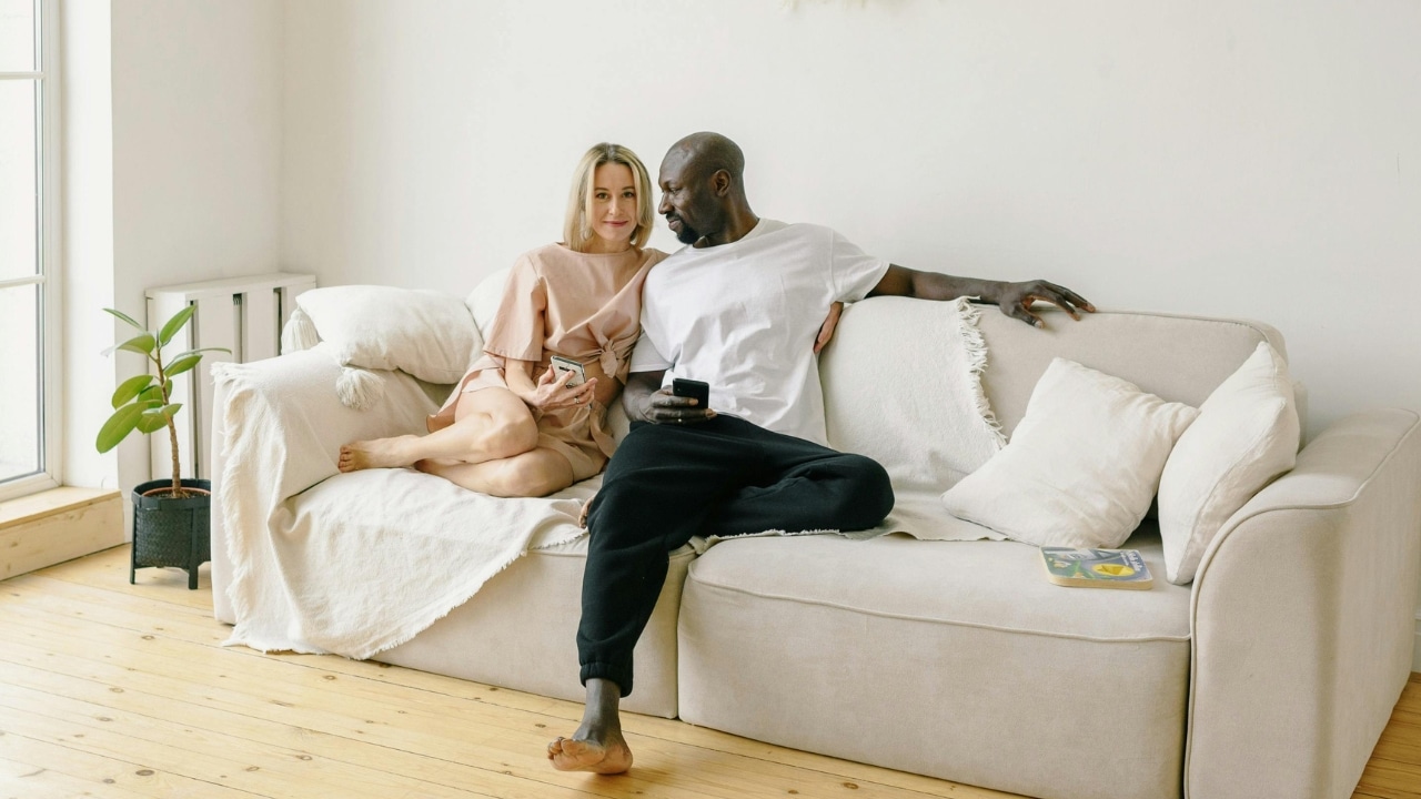 A man and woman sit on a light-colored sofa while holding their own mobile phones.