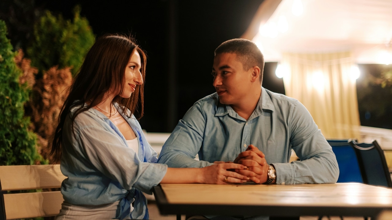 A man and a woman sit at a table outdoors at night holding hands.