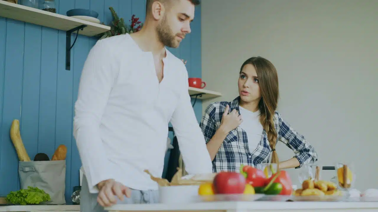A woman in a plaid shirt speaks to a man standing near a kitchen counter.