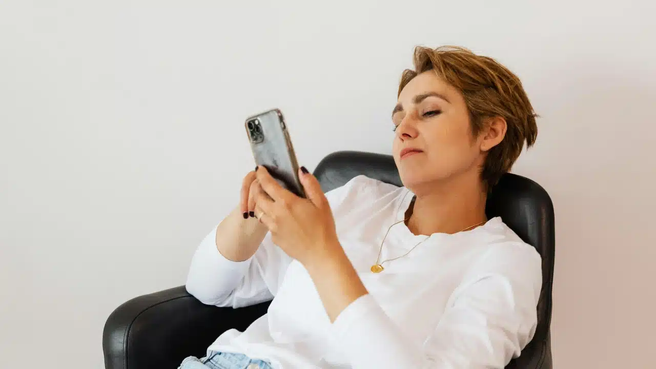 A woman with short hair reclines in a black leather chair while looking at a smartphone.