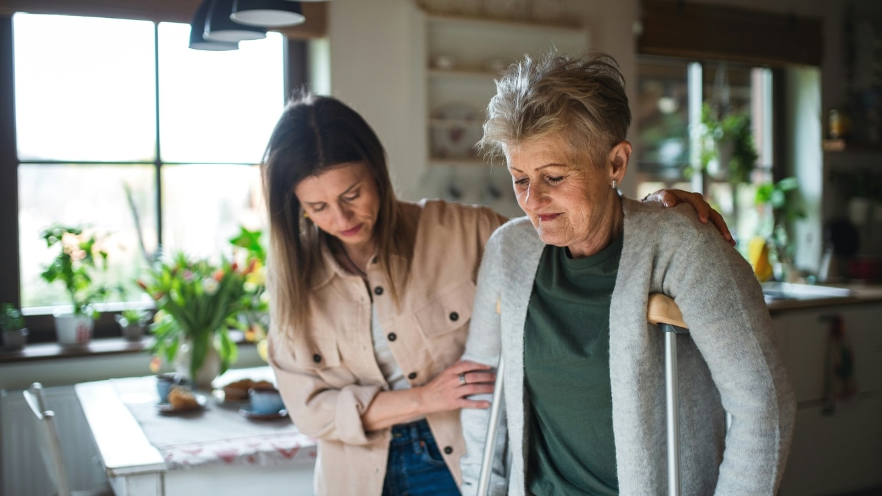 A younger woman assists an older woman using crutches to walk through a bright kitchen area.