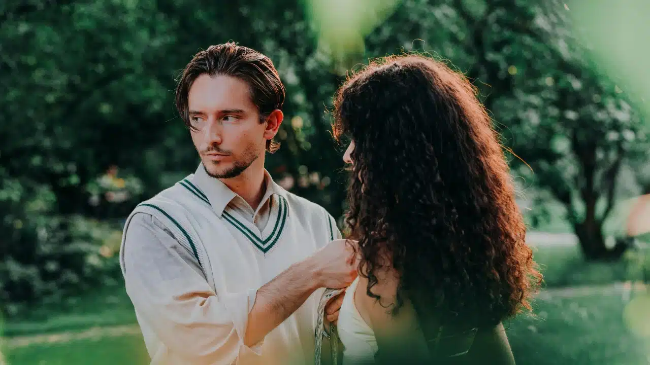 A man in a sweater vest looks over his shoulder while standing behind a curly-haired woman.
