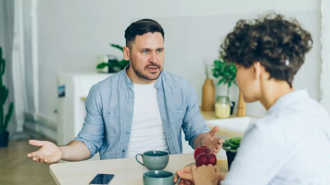 A man with an open-handed gesture speaks to a person at a wooden table.