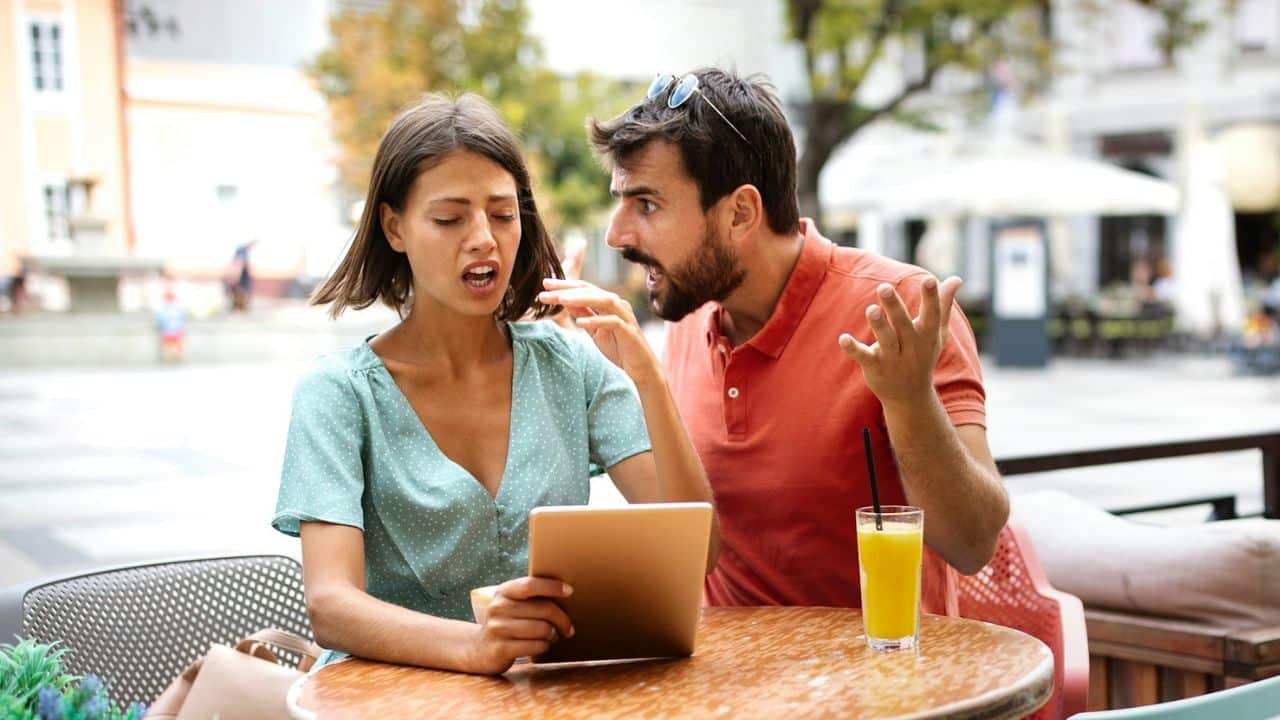 A man in an orange shirt gestures emphatically toward a woman holding a digital tablet.