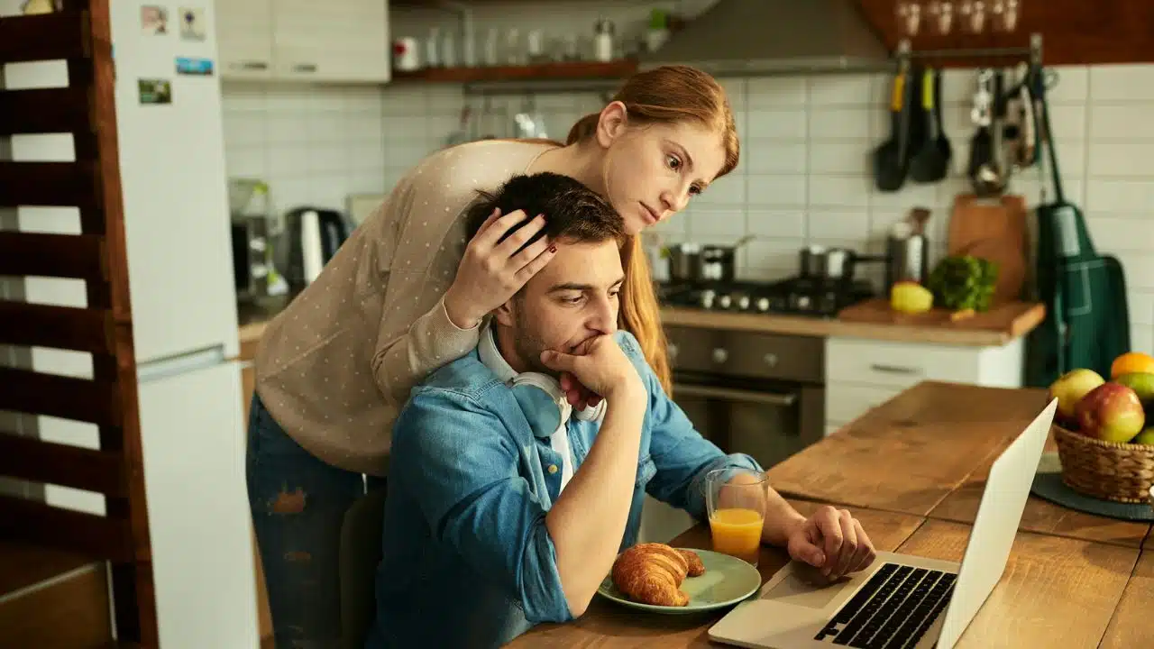 A woman leans over a man’s shoulder while he looks at a laptop in a kitchen.