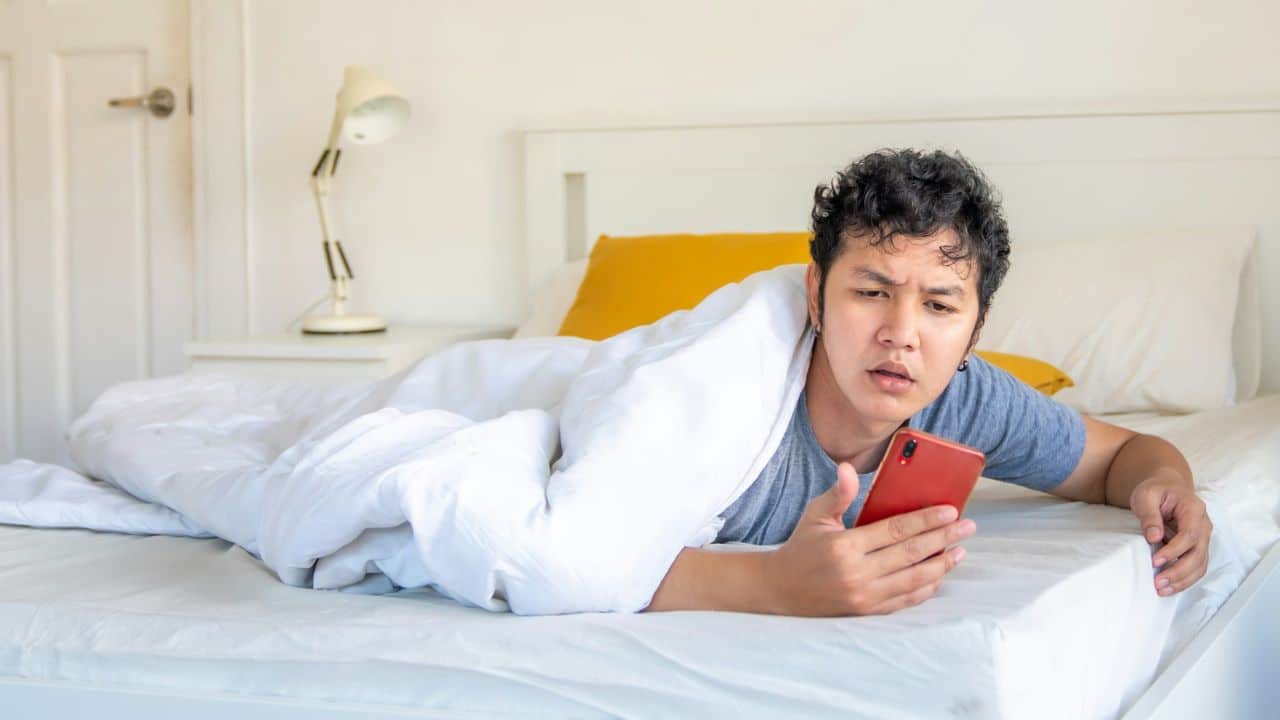 A man lies in bed under a white blanket while looking at a red smartphone.