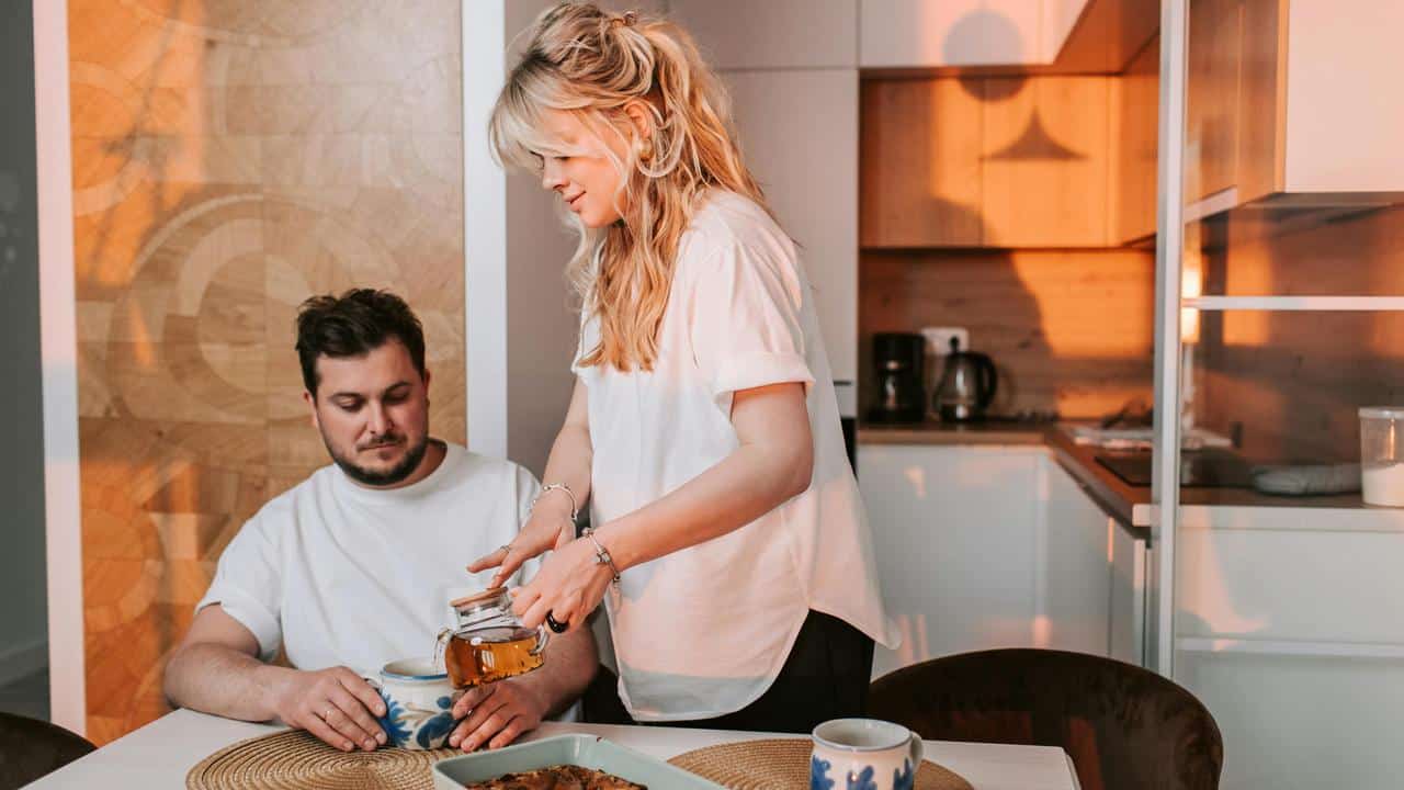 A woman pours tea for a man sitting at a table in a kitchen.