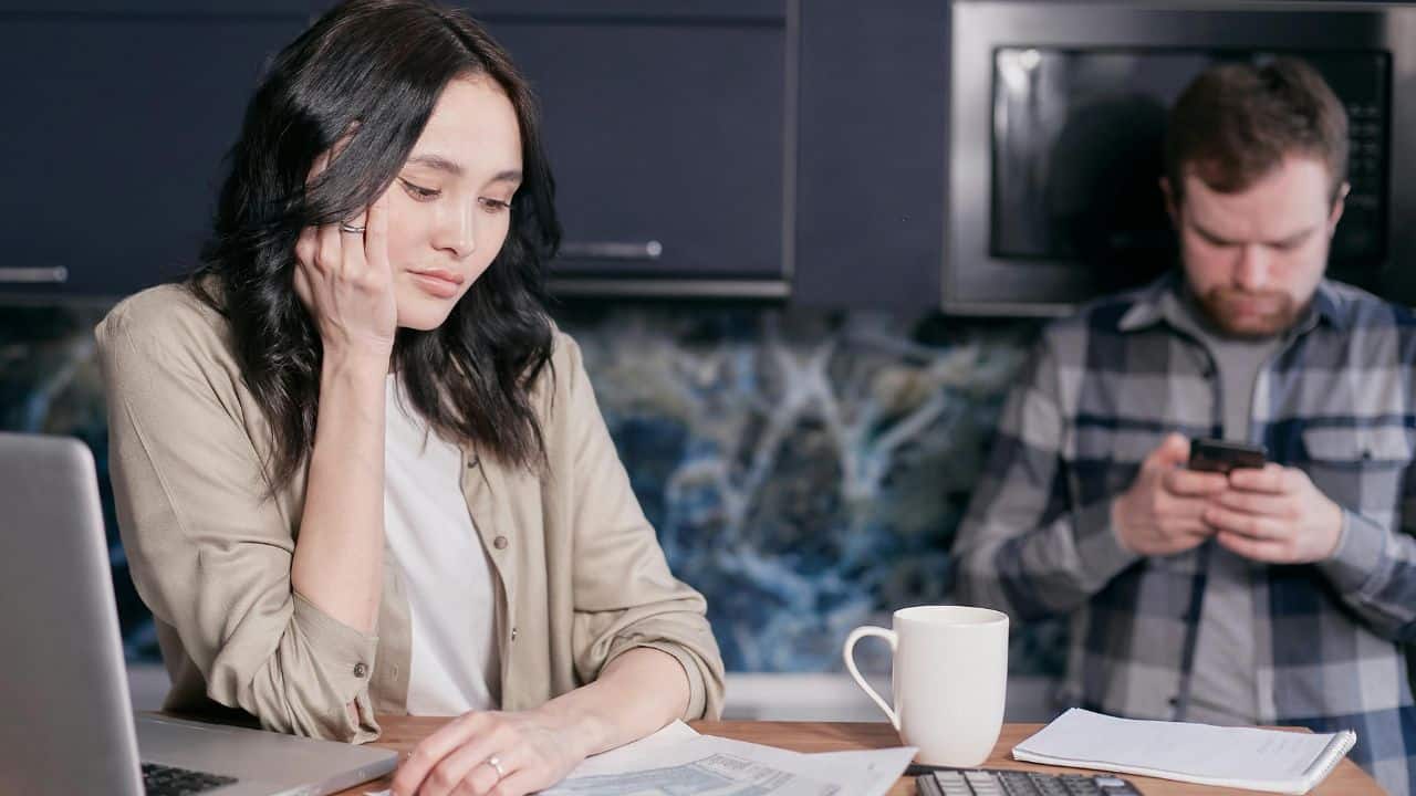 A woman sits at a table with papers while a man uses his phone behind her.