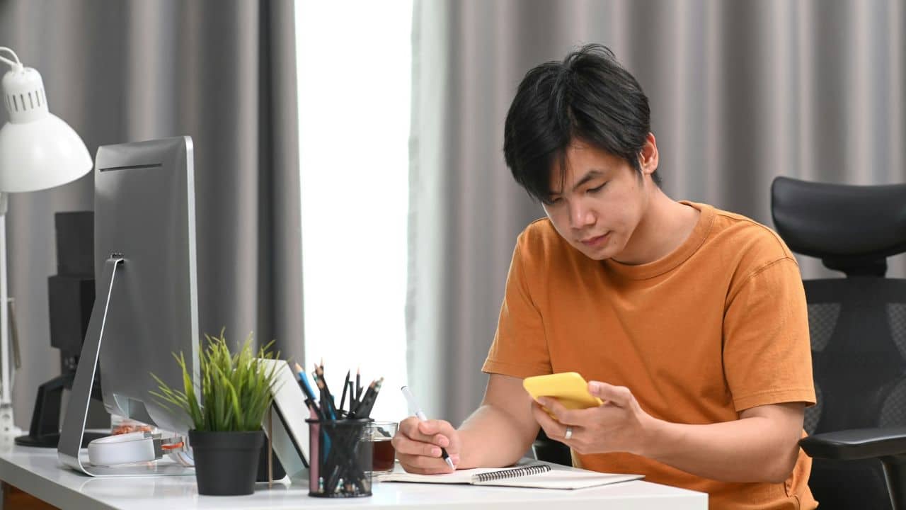 A man in an orange shirt holds a yellow phone while writing in a notebook.