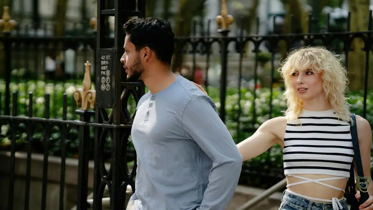 A woman in a striped top reaches out to touch a man’s shoulder outdoors.